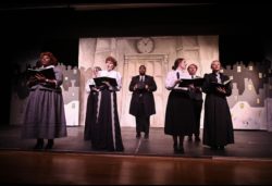Theatre still photo of a group of people singing wearing Victorian clothing.